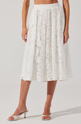 A woman is wearing a white, ankle-length skirt made of lace with intricate, floral and geometric patterns. The skirt has an elastic waistband and a flowy, layered appearance. She is also wearing white open-toed high heels with ankle straps.